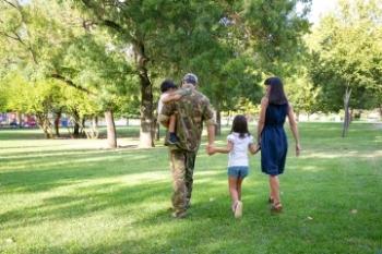 Military family in park with Dad in fatigues holding child, holding hands with wife and daughter. Banner