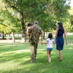 Military family in park with Dad in fatigues holding child, holding hands with wife and daughter.